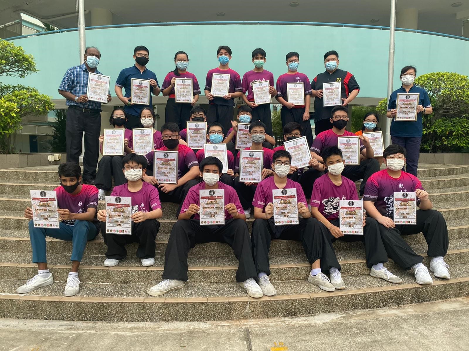 Group of masked students and instructors seated on steps outdoors holding certificates and posing together after a lion dance achievement or competition.